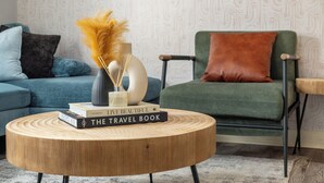 Una mesa redonda de madera con libros, un florero con flores secas y un sillón verde con un cojín marrón.