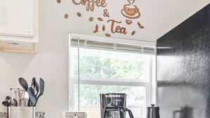 Una cocina con un cartel de café y té, una cafetera, un tostador y estantes con frascos y utensilios de cocina.
