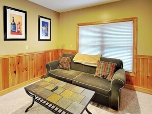 A living room with a wooden paneled wall, a green sofa with plaid cushions, a tiled coffee table, and a window with blinds. - Beautiful Day by Taylor Made Deep Creek (McHenry)
