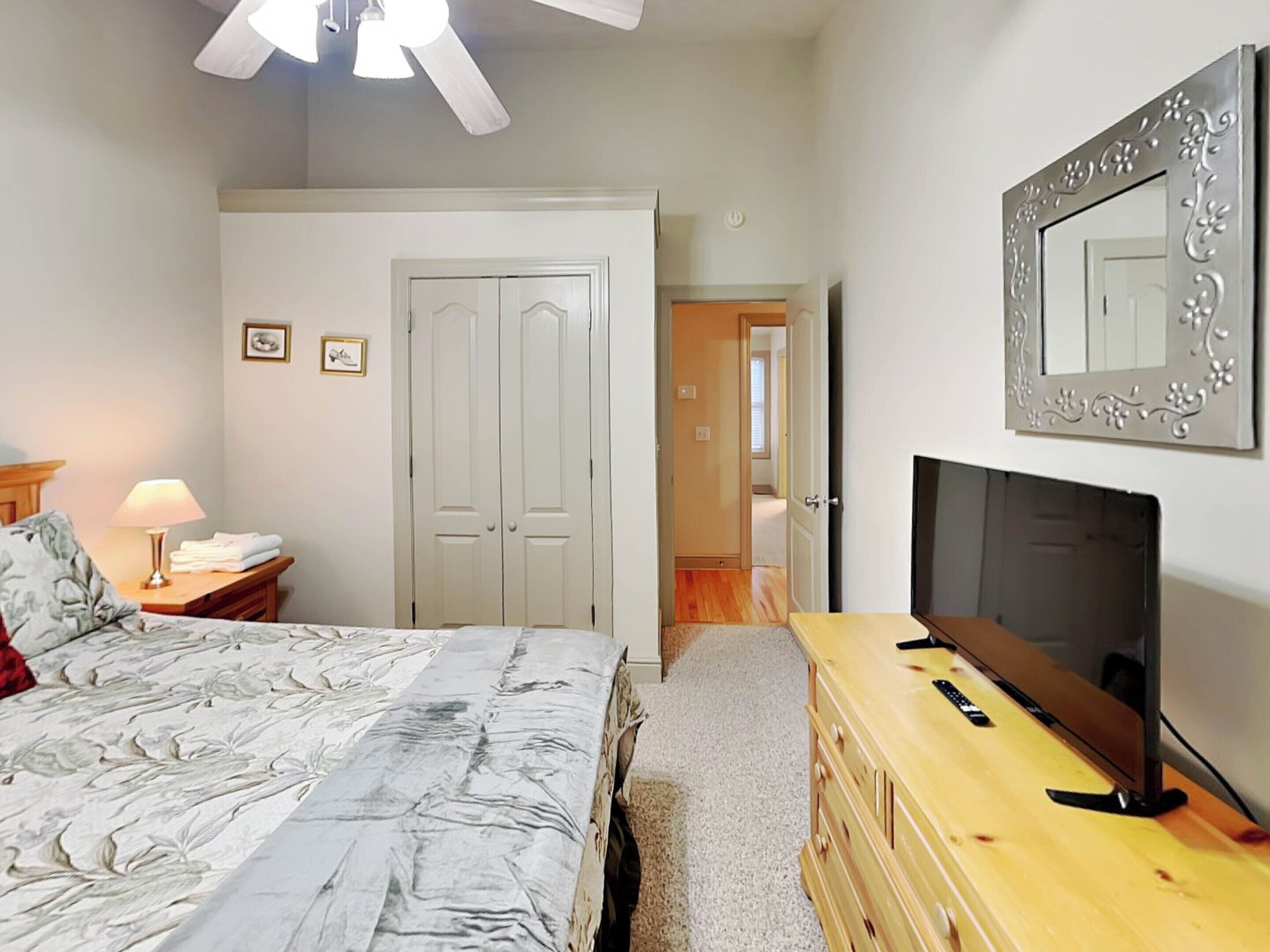 A bedroom with a bed, a dresser, a TV, a ceiling fan, and a door leading to another room.