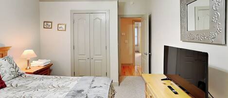 A bedroom with a bed, a dresser, a TV, a ceiling fan, and a door leading to another room.