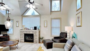 A spacious living room with a stone fireplace, a ceiling fan, and large windows. - Beautiful Day by Taylor Made Deep Creek (McHenry)