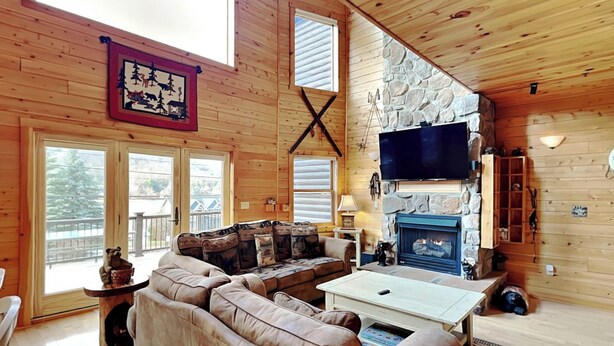 A cozy cabin interior with a stone fireplace, wooden walls, and a large window overlooking a deck. - Cedar Shores Villa 10 by Taylor Made Deep Creek (McHenry)