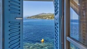 A view from a window with blue shutters, overlooking a calm sea and a distant coastline with buildings and hills. - Maison de pêcheur dans le Cap Corse (Erbalunga)