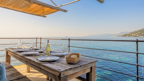 A wooden table set for dining with plates, glasses, and a bottle, overlooking a calm sea with distant land. - Maison de pêcheur dans le Cap Corse (Erbalunga)