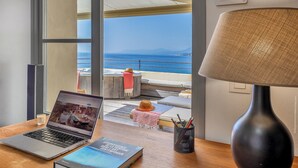 A laptop on a wooden table with a book, a lamp, and a view of the sea through large windows. - Maison de pêcheur dans le Cap Corse (Erbalunga)