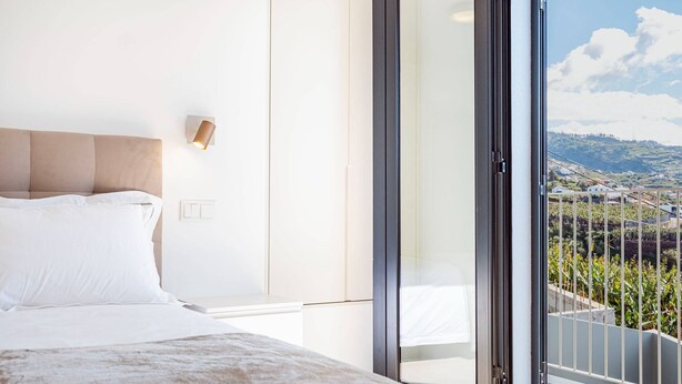 Une chambre dotée d’un grand lit, d’une table de chevet, d’une lampe fixée au mur et d’une porte-fenêtre coulissante donnant sur un balcon avec vue sur un paysage vallonné.