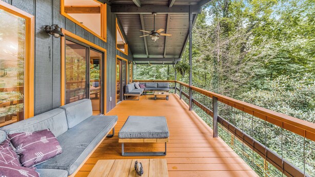 A covered outdoor deck with a sofa, ottoman, and seating area, overlooking a forested landscape.
