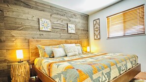 A bedroom with a wooden headboard, a patterned bedspread, a wooden floor, a ceiling fan, and a window with blinds. - Rustic charm meets comfort—king bed, AC, patio, walk 5 mins Canyon Rd, and Plaza (Santa Fe)