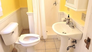 A bathroom with a toilet, sink, shower, and a mirror above the sink. - Dollhouse #4 at Portage Point Resort (Onekama)