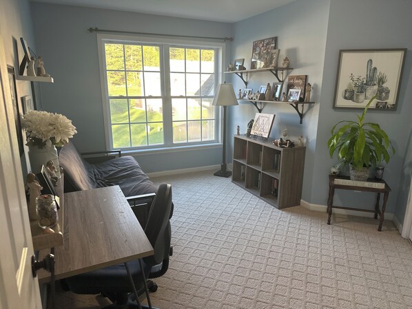 A living room with a large window, a sofa, a desk with a lamp, shelves with framed photos, and a potted plant.
