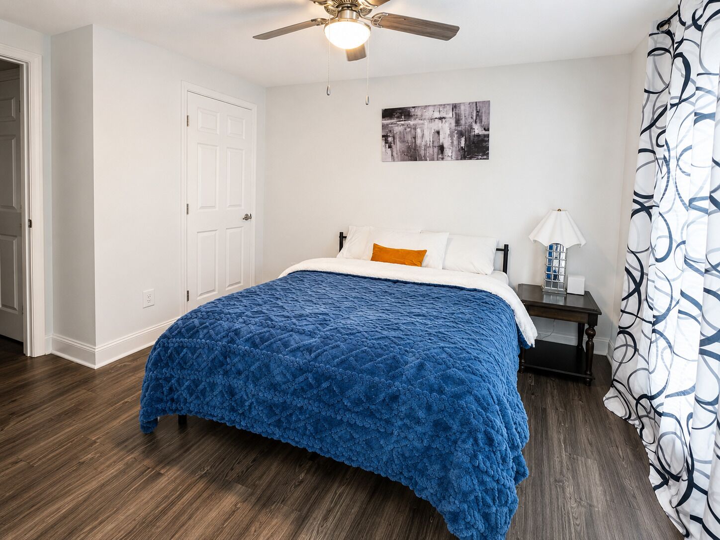 A bedroom with a bed, a nightstand, a ceiling fan, a door, and a wall-mounted artwork.