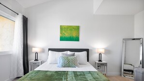 A neatly arranged hotel room with a large bed, two bedside tables with lamps, a green throw blanket, a green abstract painting on the wall, and a large mirror. - Connect With Nature at This Beautiful Urban Farm (Mesa)