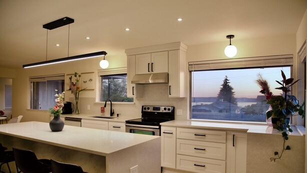 Una cocina moderna con una isla central, muebles blancos y un amplio ventanal con vistas a árboles y al atardecer.