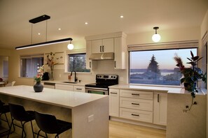 Una cocina moderna con una isla central, muebles blancos y un amplio ventanal con vistas a árboles y al atardecer.