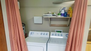 Une buanderie équipée de deux machines à laver blanches, d’une étagère contenant des produits de nettoyage et de rideaux roses.