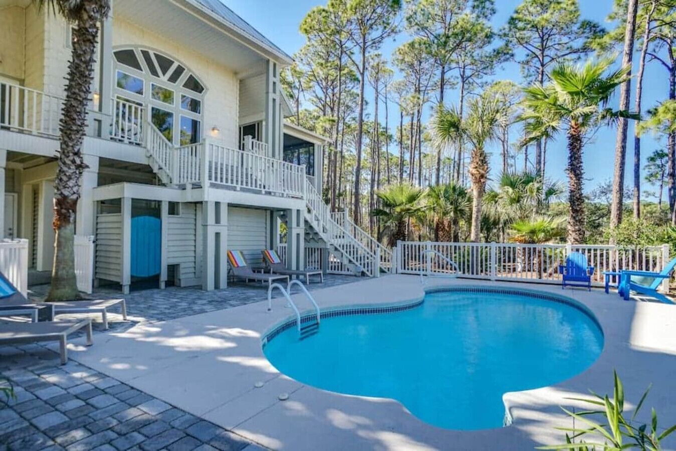 A pool area with a curved pool, a staircase leading to a balcony, and a view of tall trees and a building with a white exterior.