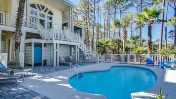 A pool area with a curved pool, a staircase leading to a balcony, and a view of tall trees and a building with a white exterior.