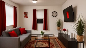 Ein Wohnzimmer mit grauem Sofa, roten Kissen, einem Couchtisch, einer Uhr, einem Fernseher und Zimmerpflanzen.