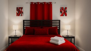 Ein Bett mit roter Bettdecke, zwei Kissen und ein weißes Handtuch auf dem Fußteil. Es gibt zwei Nachttische mit Lampen.