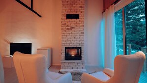 A modern living room with a brick fireplace, two white armchairs, a TV, and a large window with a view of trees. - Abant Luna (Bolu)
