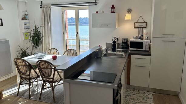 Eine moderne Küche mit einer großen Kochinsel, ein Essbereich mit Korbstühlen und Blick auf das Meer durch das Fenster.