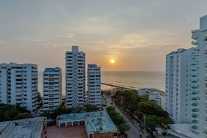 A coastal cityscape with high-rise buildings, a sunset over the ocean, and a clear view of the beach.