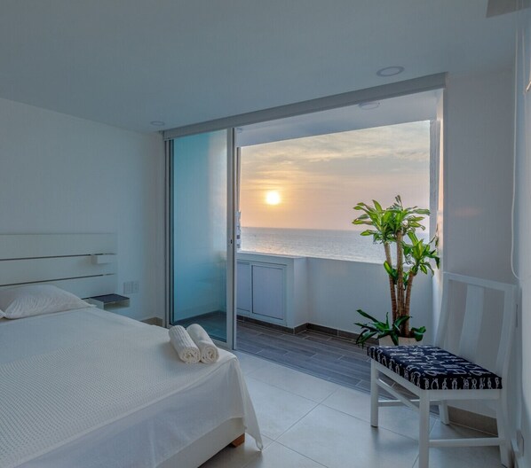 A bedroom with a bed, a chair, a plant, and a view of the sea.