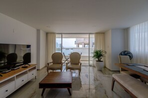 A modern living room with a flat-screen TV, two armchairs, a coffee table, a dining table, and a potted plant.