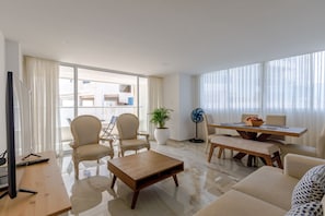 A modern living room with a flat-screen TV, a wooden coffee table, two armchairs, a dining table with chairs, a potted plant, and a standing fan.