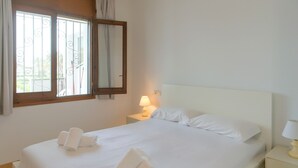Un letto rifatto con lenzuola e asciugamani bianchi, una lampada da comodino e una finestra con inferriate.