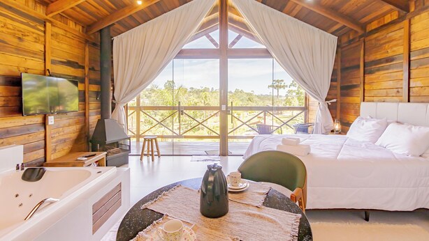 A cozy cabin with a large bed, a sitting area with a table, and a TV mounted on the wall. - VILLA ESTORIL (Campina Grande Do Sul)
