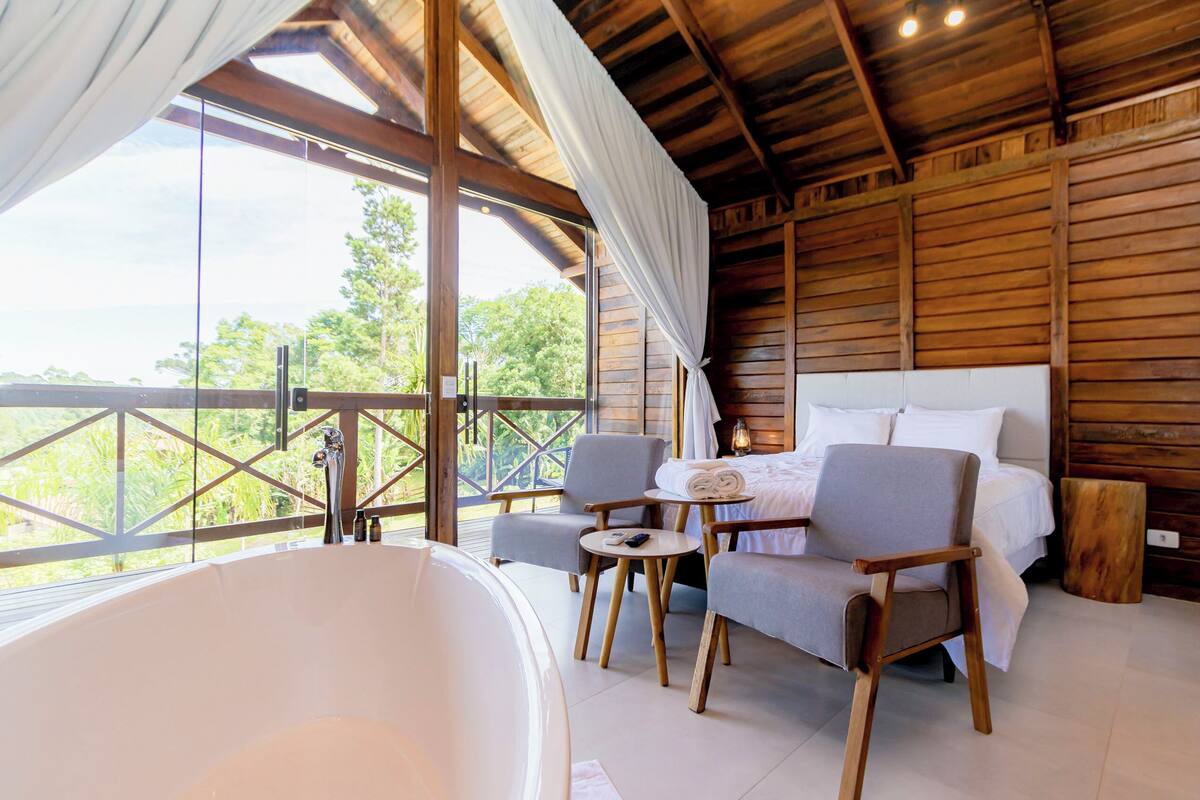 A wooden cabin with a large window, a bathtub, a bed, and two chairs.