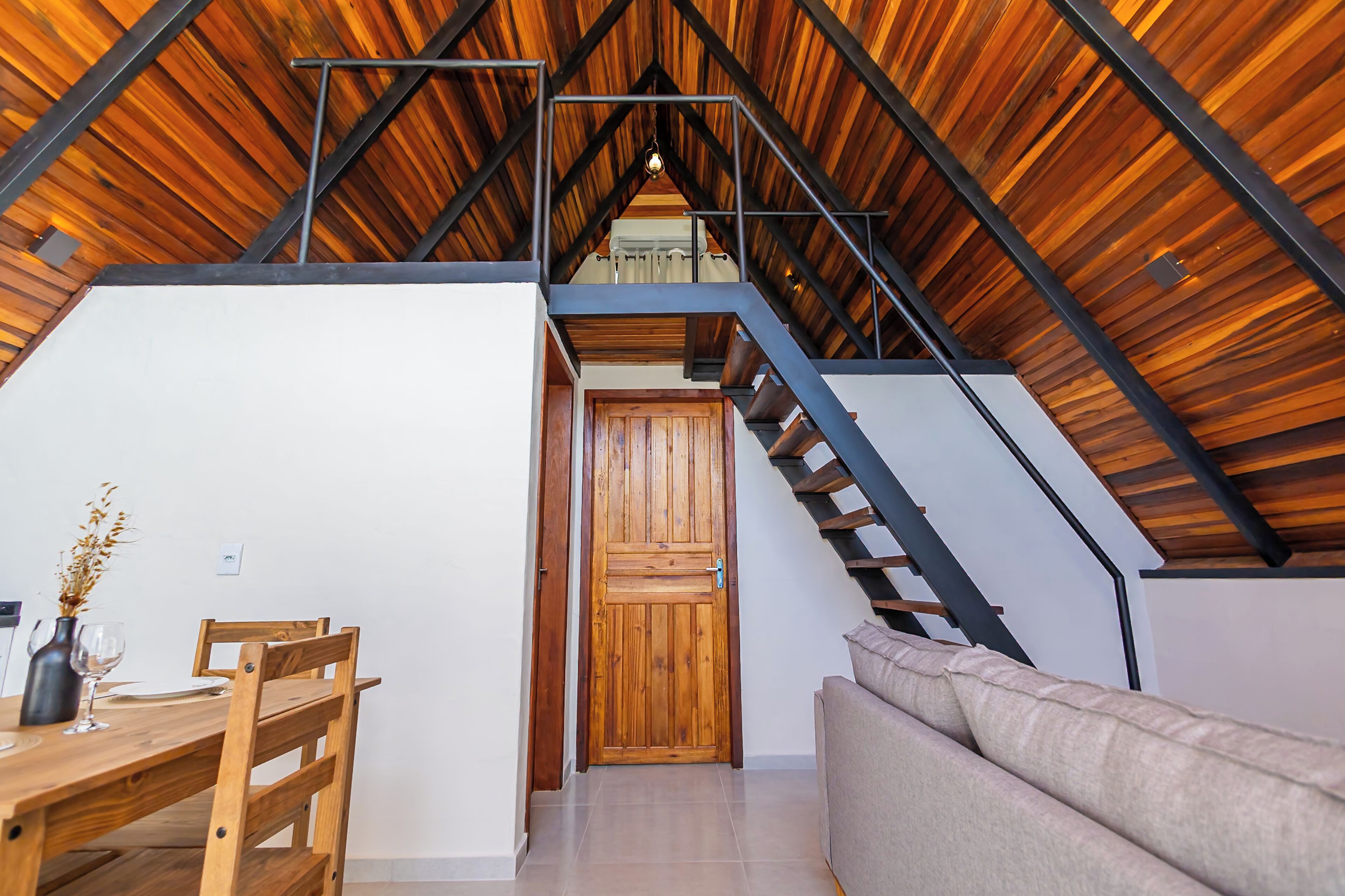 A loft-style room with a wooden ceiling, a staircase, a dining area with a table and chairs, and a sofa.