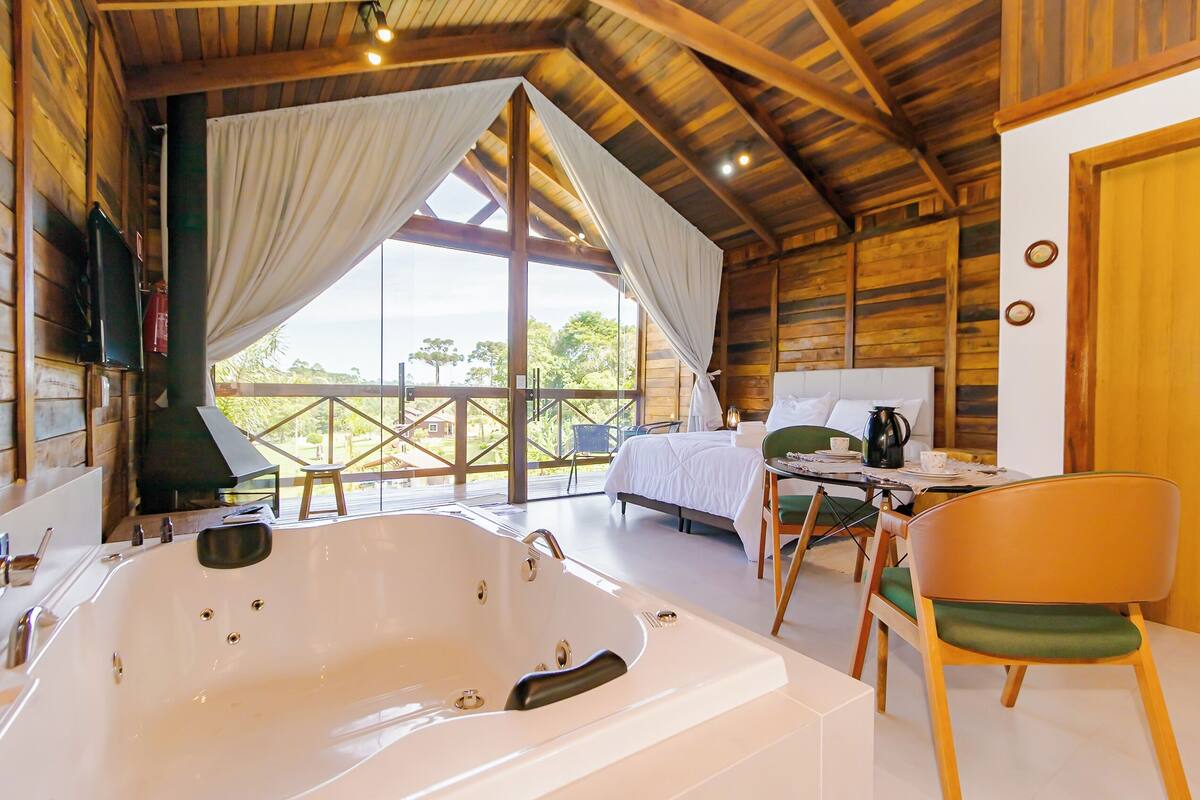 A room with a large bathtub, a bed, a dining table with chairs, and a view through large windows.
