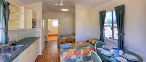 A compact room with a kitchenette, a bed with a colorful quilt, a dining table with plates and glasses, and a window with curtains.