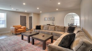 Amplia sala de estar con un sofá modular grande, dos sillones y una mesa de centro. Se observa un comedor a través de un arco.
