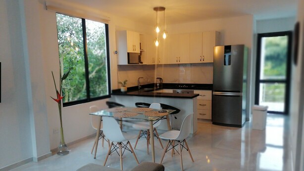 Una cocina moderna con armarios blancos, un refrigerador de acero inoxidable y un comedor con mesa de vidrio y sillas blancas.