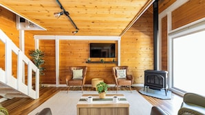 Interior acogedor de una cabaña con techo de madera, chimenea, televisor montado en la pared y escalera que sube a un nivel superior.
