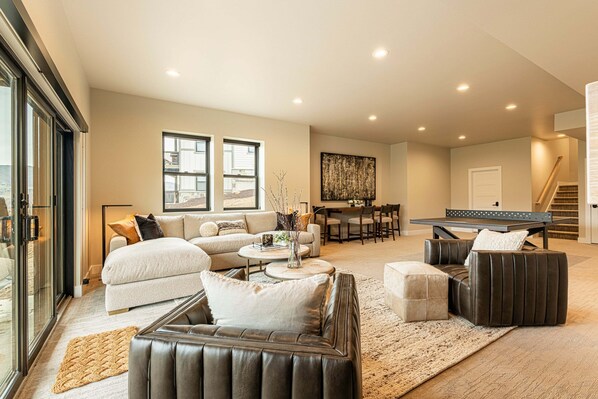 A spacious living room with a sofa, coffee table, and ottoman, a dining area with chairs, a piano, and a staircase.