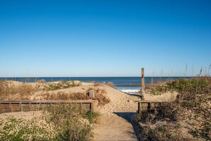 Een zandpad langs het strand dat uitkomt bij de oceaan, met houten constructies en begroeiing.