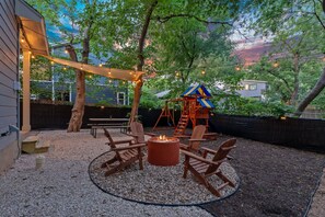 A backyard with a fire pit, wooden chairs, a swing set, and string lights.