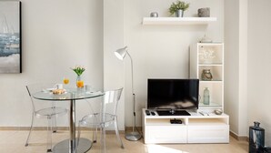 Ein modernes Wohnzimmer mit einem Glastisch, klaren Stühlen, einem Flachbildfernseher, einer Stehlampe und einer weißen Unterhaltungsanlage mit Regalen.