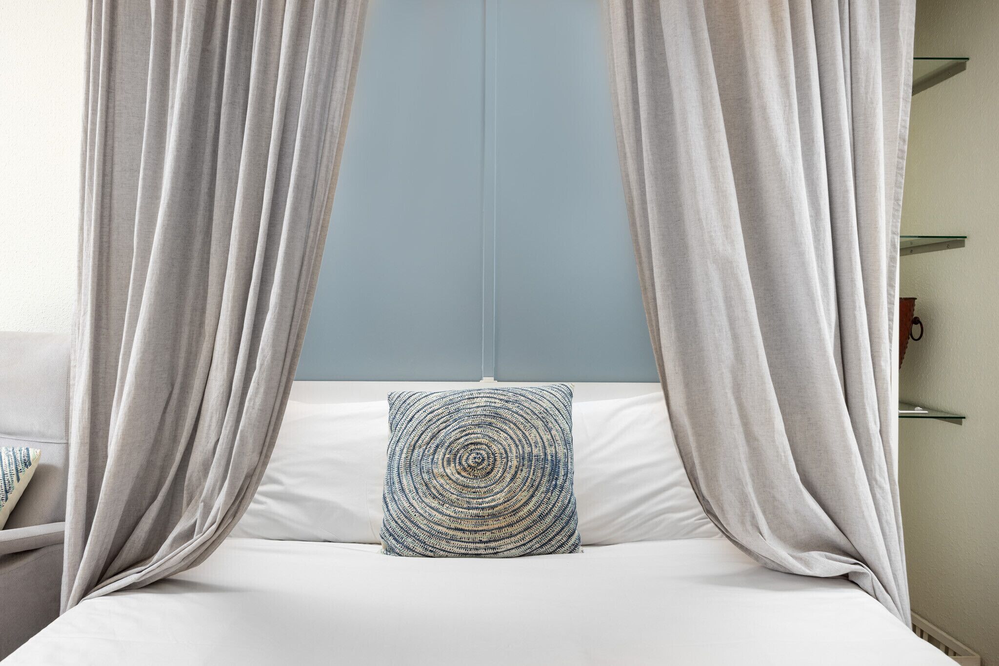 A bed with white bedding and a circular patterned pillow, draped with light-colored curtains.