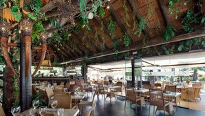 Un restaurant en plein air avec des tables et des chaises en bois, entouré de verdure et doté d’un toit de chaume.