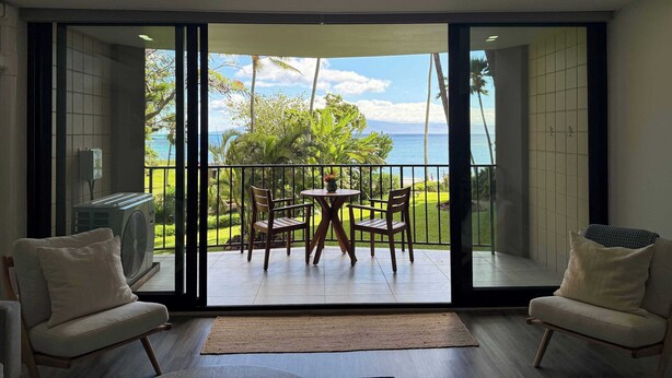 A room with a sliding glass door leading to a balcony with a view of the ocean and palm trees. There is a ceiling fan, a table with chairs, and two armchairs. - Valle Isle (Lahaina)
