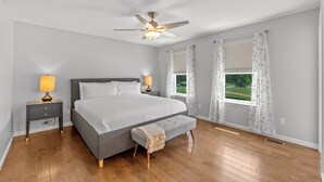 A bedroom with a bed, bedside lamps, a ceiling fan, a window with curtains, and a view of greenery outside. - Spacious 4BR w/ Garage Near SLU & Hospital | Park-Adjacent | STL (St. Louis)