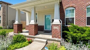 A two-story brick house with a white porch and a blue door. - Spacious 4BR w/ Garage Near SLU & Hospital | Park-Adjacent | STL (St. Louis)