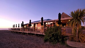 Ein Gebäude direkt am Strand mit einer Holzterrasse, die sich über den Sand erstreckt, mit mehreren Sonnenschirmen und einer Palme.
