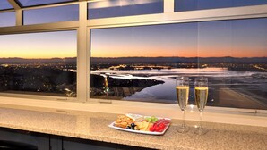 Ein Panoramablick vom Fenster eines Hotelzimmers auf eine Stadtlandschaft im Abenddämmerlicht, mit einem Fluss, einer Teller mit Snacks und zwei Sektgläsern auf der Anrichte.